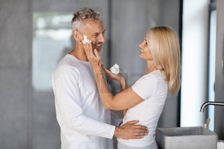 Photo for Happy Mature Couple Spending Time In Bathroom Together, Caring Wife Applying Shaving Foam On Husbands Face And Laughing, Romantic Senior Spouses Making Beauty Routine At Home, Free Space - Royalty Free Image
