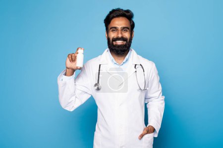 Foto de Médico indio de mediana edad positivo en uniforme sosteniendo frasco de medicamentos, de pie sobre fondo azul. Pastillas para el cuidado de la salud, consejos de terapeuta para el tratamiento - Imagen libre de derechos