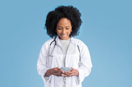 Téléchargez les photos : Heureuse jeune femme noire médecin thérapeute heureuse en manteau blanc tapant sur smartphone, bavardant, isolée sur fond de studio bleu. App pour la médecine, soins de santé avec gadget, message - en image libre de droit