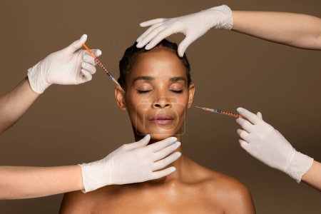 Photo for Attractive african american middle aged woman getting facial injections by two cosmetologists, beautician hands in gloves giving anti aging serum shots, brown studio background - Royalty Free Image