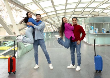 Photo for Travel With Friends. Two Women Jumping And Hugging Their Husbands, Posing Together And Having Fun At Modern Airport Interior, Smiling At Camera. Group Of Young Passangers Celebrating Vacation - Royalty Free Image