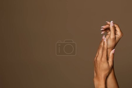 Téléchargez les photos : Vue rapprochée des mains féminines afro-américaines avec manucure, femme touchant une peau douce après un traitement spa et appliquant de la crème, fond de studio brun, espace libre - en image libre de droit