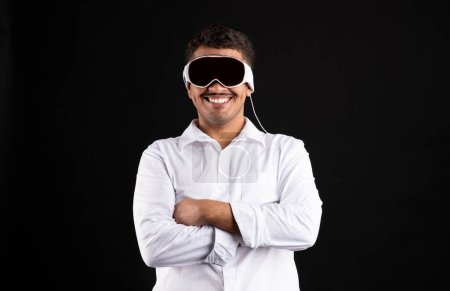Foto de Emocionado hombre brasileño con gafas Visión Pro VR, de pie con los brazos cruzados y sonriendo, utilizando gadget moderno para el entretenimiento en fondo negro estudio - Imagen libre de derechos