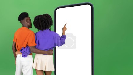 Foto de Millennial africano americano pareja señalar dedo en el teléfono inteligente grande con pantalla en blanco, aislado en fondo de estudio verde, espalda. Aplicación, blog, sitio web en venta, banca, redes sociales, anuncio y oferta - Imagen libre de derechos