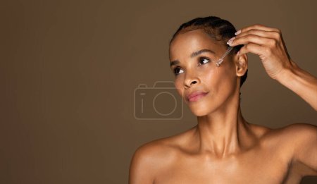 Téléchargez les photos : Belle femme afro-américaine d'âge moyen avec une peau parfaite appliquant du sérum sur son visage, isolée sur fond brun, espace de copie. Soins de beauté, cosmétiques et soins anti-âge spa - en image libre de droit