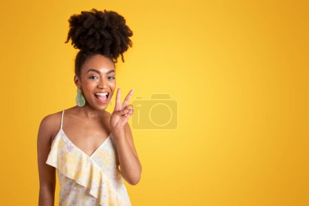 Photo for Glad young black woman in dress with open mouth making peace sign with hand, isolated on yellow background, studio. Lifestyle recommendation, good news gesture, fun emotions, ad and offer - Royalty Free Image