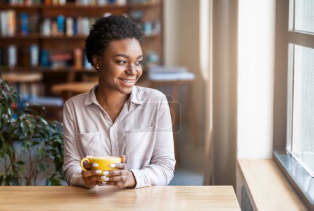 Téléchargez les photos : Souriant jolie jeune femme noire aux cheveux courts en tenue décontractée intelligente avec une tasse de café frais assis seul à table dans un café, regardant par la fenêtre et souriant, rêvant de bonne journée - en image libre de droit
