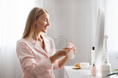 Téléchargez les photos : Belle dame mature appliquant une lotion démaquillante sur un coton pour nettoyer le visage, souriante belle femme d'âge moyen faisant une routine quotidienne de soins de la peau à la maison, bénéficiant de soins de beauté - en image libre de droit