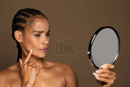 Téléchargez les photos : Frustré afro-américaine d'âge moyen tenant miroir et regardant bouton sur son visage, souffrant de problèmes de peau ou remarquant des changements d'âge, debout sur fond de studio brun - en image libre de droit