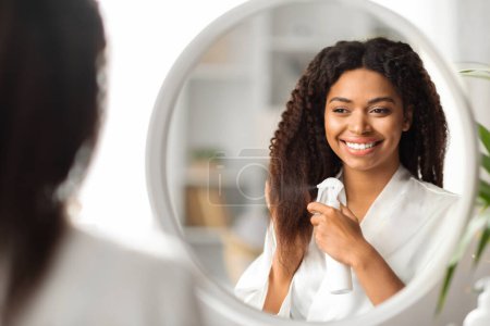 Foto de Hermosa mujer negra aplicando spray hidratante en el cabello mientras está parada cerca del espejo en casa, dama afroamericana disfrutando de nuevos cosméticos de peinado, sonriendo a su reflejo, enfoque selectivo - Imagen libre de derechos