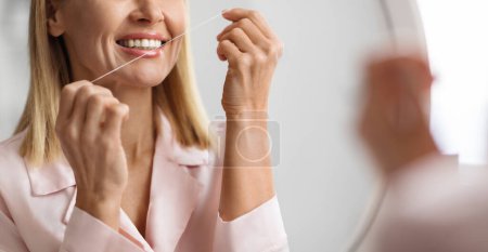 Zugeschnittene Aufnahme einer lächelnden Frau mittleren Alters mit Zahnseide zu Hause, glückliche reife Frauen, die Mundpflege machen, neben Spiegel im Badezimmer stehen, Zähne reinigen, tägliche Hygiene-Routine, Nahaufnahme