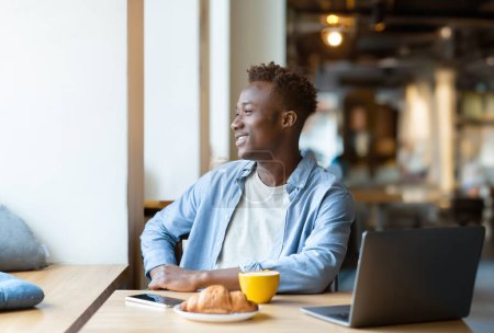 Téléchargez les photos : Souriant jeune homme noir à l'aide d'un ordinateur portable dans un café confortable, prendre une boisson chaude avec croissant, regarder par la fenêtre. Beau Afro-Américain étudiant ou travaillant en ligne dans un café - en image libre de droit