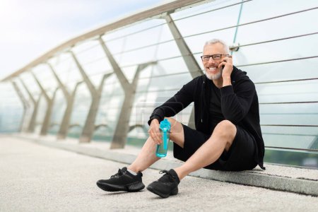 Photo for Positive well-fit senior sportsman in black sportswear sitting on bridge, holding bottle with water, have phone conversation while resting after workout outdoor, looking at copy space - Royalty Free Image