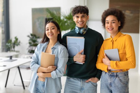 Photo for Happy three multiracial students posing in coworking space, cheerful multiethnic young people with workbooks standing in classroom and smiling, copy space - Royalty Free Image