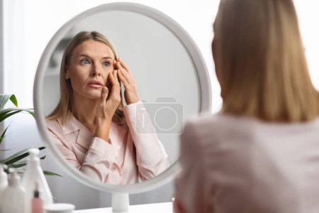 Aging Signs. Upset mature woman looking at wrinkles around her eyes while standing near mirror at home, beautiful middle aged lady inspecting face, feeling stressed, selective focus