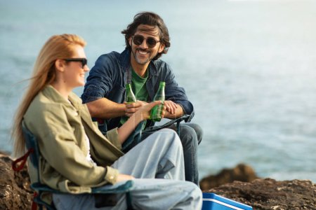 Photo for Smiling Man And Woman Relaxing Together On Rocks At Ocean Shore, Young Happy Couple Chatting And Drinking Soda, Sitting In Camping Chairs Outside, Having Fun On Outdoor Date, Free Space - Royalty Free Image