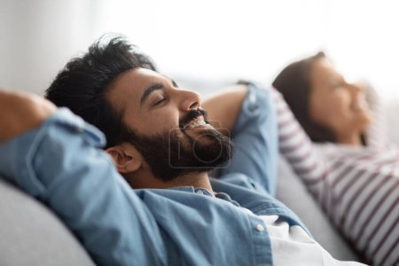 Photo for Relaxation concept. Satisfied relaxed beautiful eastern young spouses husband and wife resting on sofa at home. Happy indian couple reclining on couch in living-room, selective focus on bearded man - Royalty Free Image