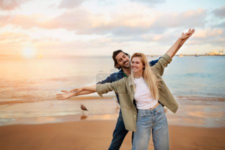 Téléchargez les photos : Jeune couple sur la plage faisant semblant de voler, écartant les mains comme des ailes, joyeux homme et femme millénaire s'amuser tout en marchant au bord de la mer, rire et profiter de la date en plein air, copier l'espace - en image libre de droit