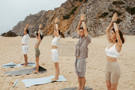 Téléchargez les photos : Groupe de jeunes hommes et femmes sportifs pratiquant le yoga sur la plage, faisant la queue et levant les mains, profitant de l'entraînement matinal sur le rivage de l'océan, copiez l'espace - en image libre de droit