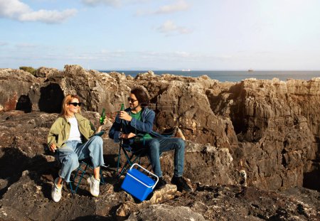 Téléchargez les photos : Joyeux jeune couple buvant du soda tout en se relaxant dans des chaises de camping sur des rochers de plage, heureux homme et femme millénaire se reposant près de l'océan, pique-nique ou rendez-vous en plein air, s'amuser ensemble, copier l'espace - en image libre de droit