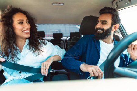 Téléchargez les photos : Jeunes épouses excitées du Moyen-Orient attachant les ceintures de sécurité, assises dans une voiture neuve à l'intérieur, se regardant et souriant. Concept d'offre de services de sécurité des passagers et d'assurance automobile - en image libre de droit