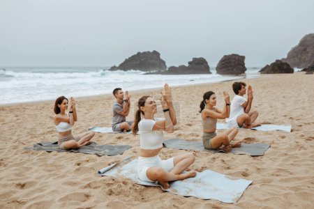 Téléchargez les photos : Groupe de femmes heureuses et l'homme faisant du yoga sur les tapis de fitness, l'entraînement le matin sur la plage de sable, les gens assis avec les jambes croisées et faire la pose namaste - en image libre de droit