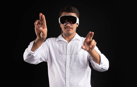 Foto de Hombre latino experimentando la realidad virtual en gafas pro vr de visión moderna, interactuando con pantallas virtuales haciendo gestos de manos y tocando la pantalla táctil invisible, fondo negro - Imagen libre de derechos