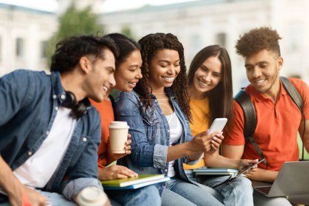 Téléchargez les photos : Loisirs du campus. Groupe multiethnique d'étudiants se relaxant ensemble à l'extérieur, heureux jeunes hommes et femmes multiculturels utilisant un smartphone ensemble, se reposant après les cours, bavardant et riant - en image libre de droit