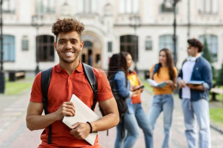 Photo for High Education Concept. Portrait of happy black male student smiling at camera while posing outdoors in campus, his classmates and university building on background, selective focus - Royalty Free Image