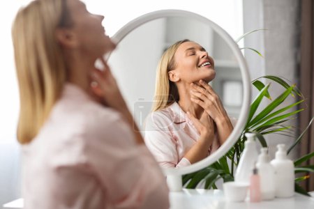 Photo for Double Chin Treatment. Beautiful Middle Aged Woman Touching Soft Smooth Skin On Neck, Attractive Mature Lady Sitting Near Mirror At Home, Enjoying Domestic Beauty Routine, Copy Space - Royalty Free Image
