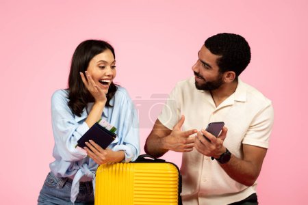 Photo for Happy cheerful young multicultural couple travellers going vacation together, carrying luggage, using cell phone, booking hotel online, carrying plane tickets and passports, pink background - Royalty Free Image