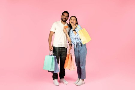 Photo for Happy cheerful beautiful young multicultural couple shopping together on pink studio background, handsome guy and pretty lady holding colorful paperbags, purchases and bank card, smiling at camera - Royalty Free Image