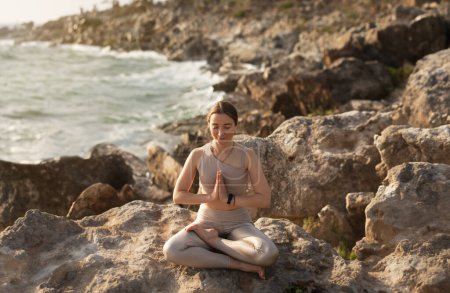 Foto de Feliz calma joven atleta europea en ropa deportiva sentarse en posición de loto, practica yoga, disfruta del entrenamiento al aire libre y ejercicios de respiración en la playa de mar. Descanso solo, cuidado de la salud, deporte y vitalidad - Imagen libre de derechos