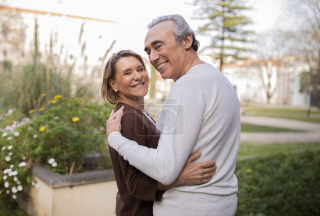 Téléchargez les photos : Joyeux couple de retraités. Épouses matures romantiques embrassant sourire à la caméra posant portant des tenues décontractées dans le parc de la ville à l'extérieur. Homme et femme jouissant de leur amour doré debout en plein air le week-end - en image libre de droit