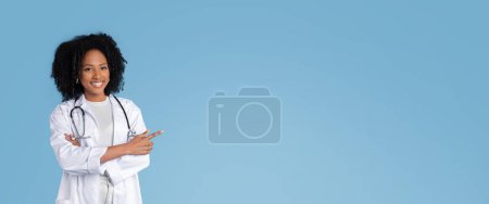 Photo for Cheerful young black woman doctor therapist in white coat pointing finger at copy space, isolated on blue studio background. Ad and offer, recommendation health care, panorama - Royalty Free Image