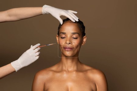 Téléchargez les photos : Belle femme afro-américaine d'âge moyen se faisant injecter le visage dans la zone des pommettes, debout sur fond de studio brun. Cosmétique beauté injections concept - en image libre de droit