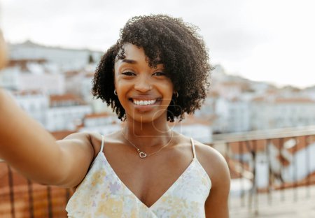 Téléchargez les photos : Une jeune femme afro-américaine souriante se promène en ville, apprécie le voyage, prend une vidéo ou un selfie au téléphone, en plein air. Appel vidéo, chat en ligne, blog de voyage, application pour les vacances, week-end et voyage seul - en image libre de droit