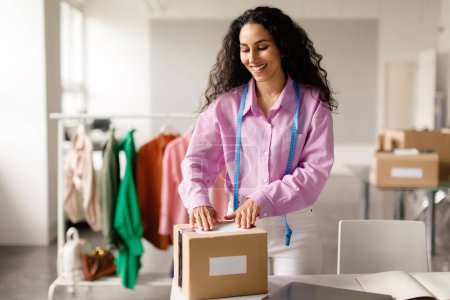 Téléchargez les photos : Happy Dressmaker femme boîte d'emballage avec commande de vêtements, bénéficiant du processus d'emballage dans la conception de mode de travail à l'intérieur du showroom moderne. Petits vêtements Commerce de détail, Commerce - en image libre de droit