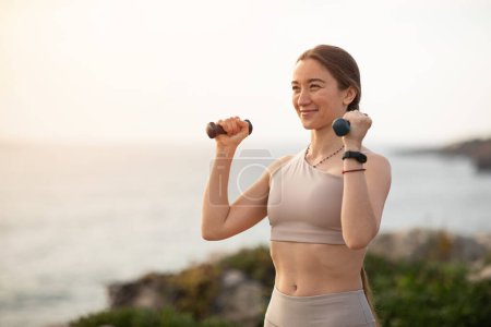 Photo for Smiling young european lady athlete in sportswear make arm exercise with dumbbells workout on sea beach. Active lifestyle and motivation, sports outdoor, weight loss and body care - Royalty Free Image