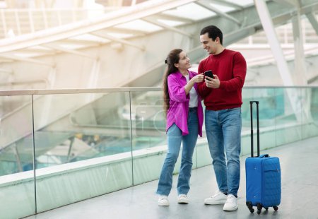 Photo for Travel App. Happy Tourists Couple Posing With Smartphone And Suitcase Standing At Modern Airport, Woman Pointing At Mobile Phone Recommending Application And Booking Tickets Online. Copy Space - Royalty Free Image