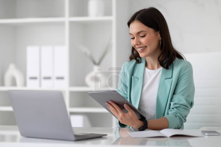 Foto de Sonriente joven mujer europea en traje en la mesa con el ordenador portátil escribiendo en la tableta en el interior de la oficina moderna. Aplicación para negocios, trabajo, estilo de vida de mujer de negocios con dispositivo, redes sociales - Imagen libre de derechos