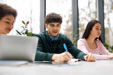 Photo for Study process concept. Multiracial students sitting at desk learning together, preparing for university exams, using laptop and taking notes, classroom or coworking space - Royalty Free Image