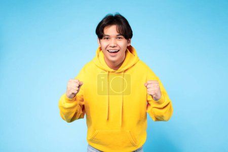 Photo for Yes, Victory Concept. Emotional Japanese teenager guy shaking fists and gesturing yes, celebrating success posing over blue studio background. Joy and celebration of teen age achievement - Royalty Free Image