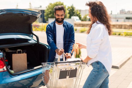 Téléchargez les photos : Jeunes conjoints du Moyen-Orient emballant des produits d'épicerie dans le coffre de la voiture à l'extérieur, debout sur le parking près du centre commercial. Ventes en supermarché - en image libre de droit