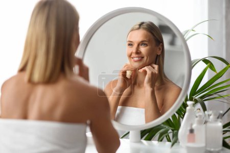 Facebuilding Concept. Beautiful Middle Aged Woman Looking In Mirror And Massaging Face, Smiling Attractive Female Touching Chin, Enjoying Anti-Aging Beauty Treatments, Selective Focus On Reflection