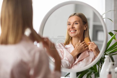 Photo for Smiling Middle Aged Blonde Lady Brushing Her Hair With Comb Near Mirror, Happy Mature Woman using Brush While Sitting At Dressing Table, Making Haircare Routine At Home, Slective Focus On Reflection - Royalty Free Image