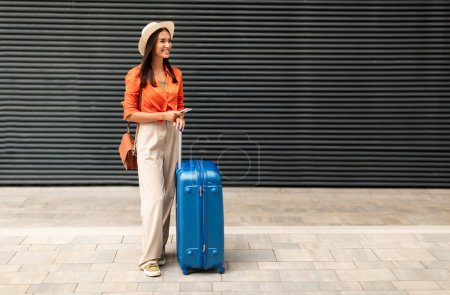 Téléchargez les photos : Happy Passenger Woman avec une valise de voyage tenant le téléphone, regardant à côté du mur avec un espace vide pour le texte, debout à l'extérieur. Tourist Woman Réservation de billets Via App. Concept de voyage vacances - en image libre de droit