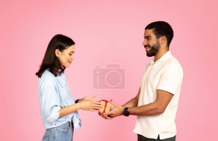 Photo for Beautiful happy young couple exchanging presents, isolated on pink studio background. Loving middle eastern guy giving gift box with red bow to his pretty girlfriend, celebrating anniversary - Royalty Free Image