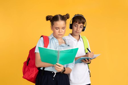 Photo for Serious teenager european boy in headphones cheating, write homework at smart girl with notebooks, isolated on yellow studio background. Friendship, childhood, education at school - Royalty Free Image