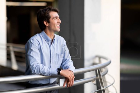 Photo for Happy handsome young man in formal outfit entrepreneur enjoying view out of office balcony, holding cell phone in his hands, looking at copy space and smiling. Business opportunities for millennials - Royalty Free Image